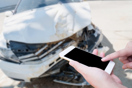 Person holding and tapping a smartphone in the foreground, with a heavily damaged car from an accident visible in the background.